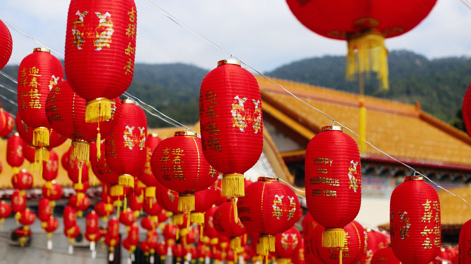 Rows of red paper lanterns with gold tassels and New Year greetings hanging in front of a temple's golden roof against a mountain backdrop.