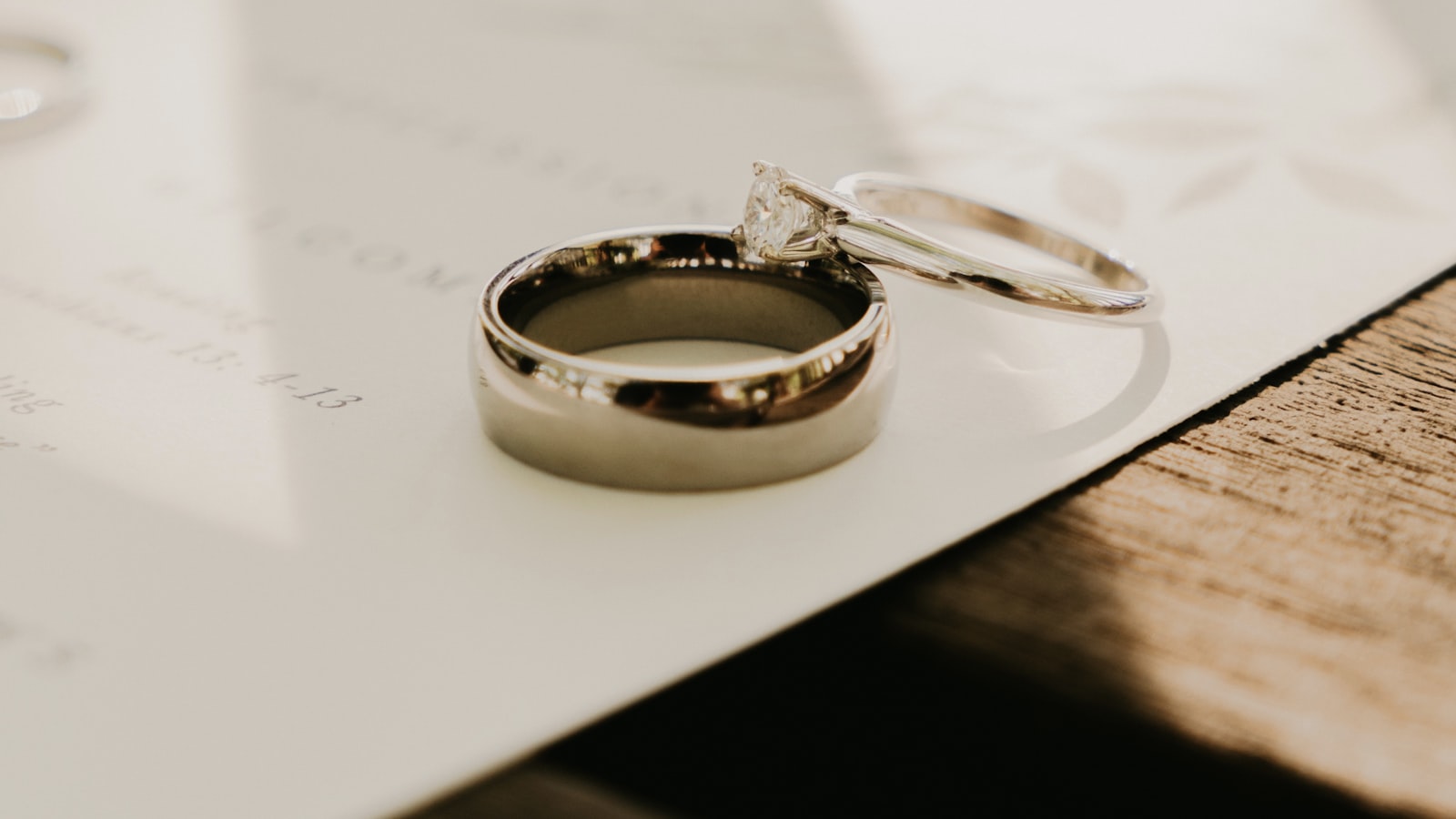Two wedding rings resting on a white invitation card, lit by warm slanted sunlight on a wooden table