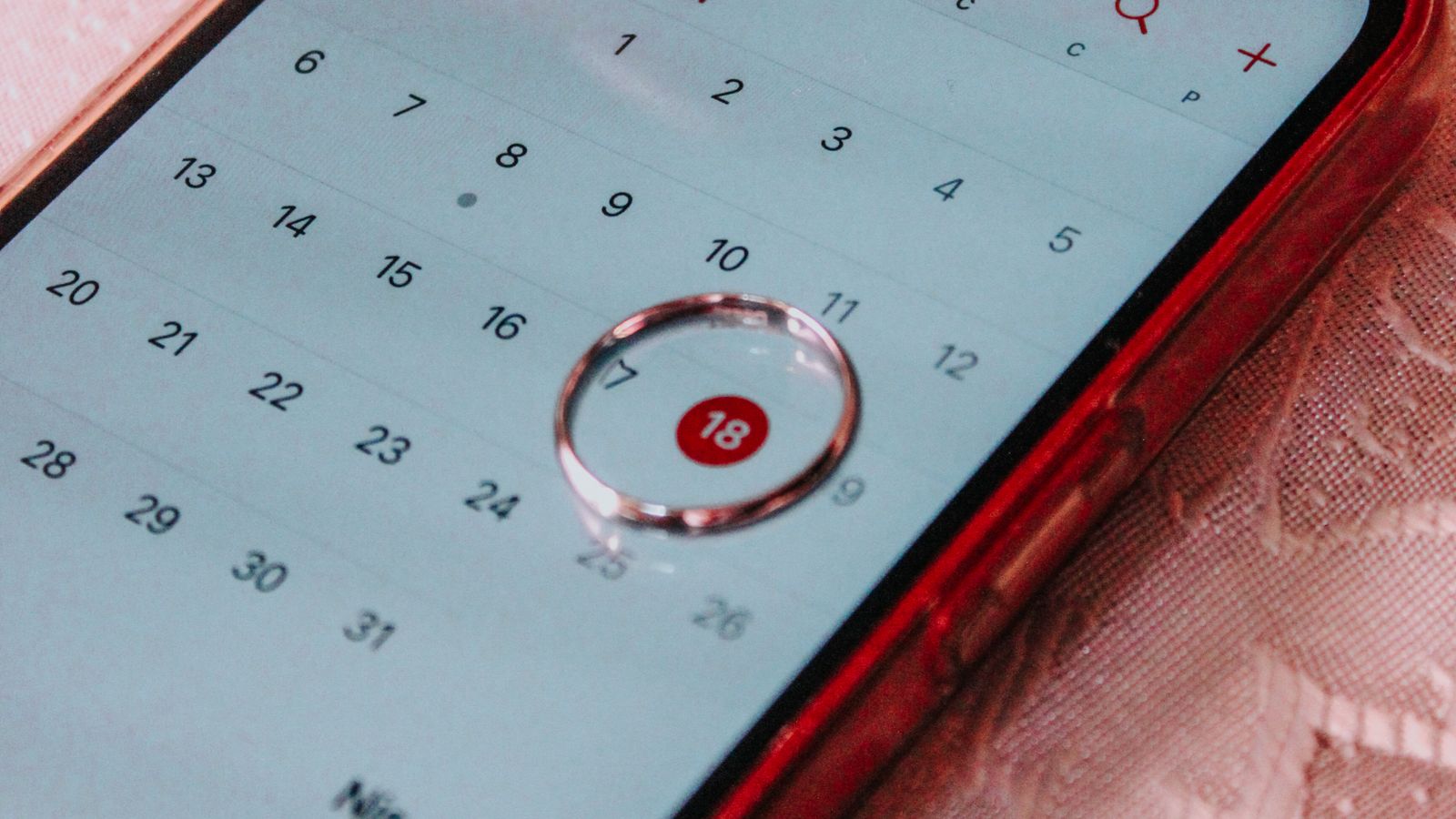 A smartphone with a red case resting on a tablecloth, its calendar app showing a specific date highlighted.