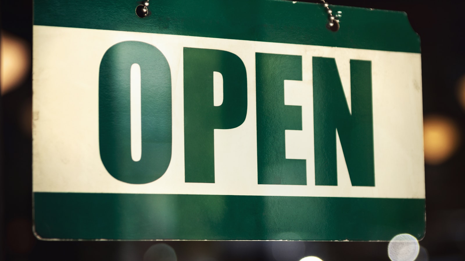 A green and white neon Open sign hanging from a chain in a shop window, with a blurred bokeh background.