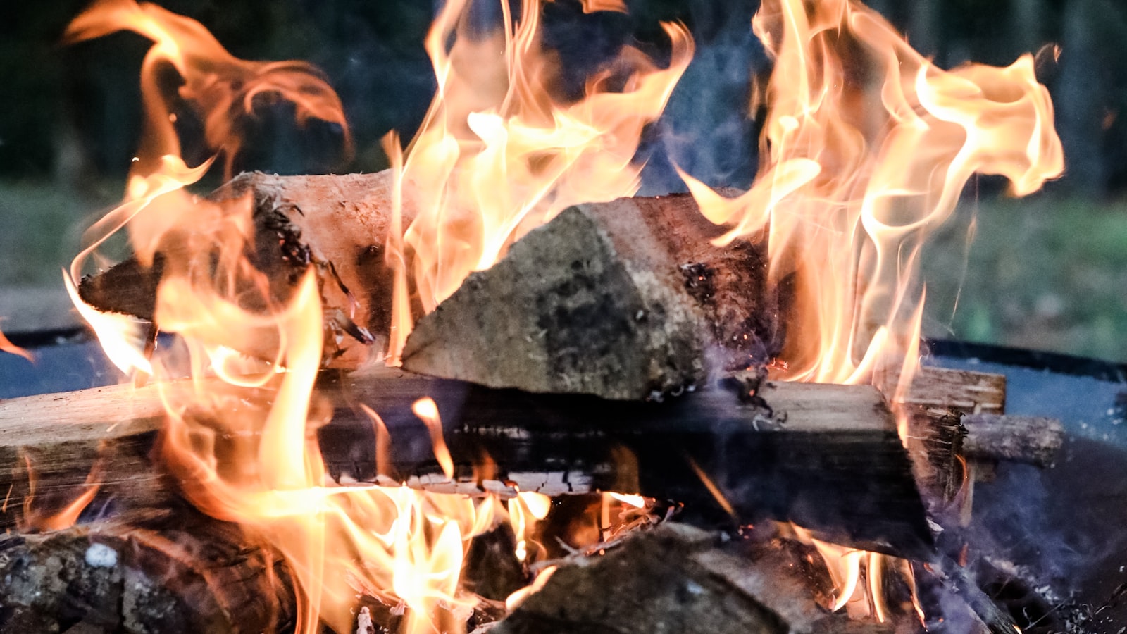 Close-up of orange flames rising around burning logs in an outdoor fire pit at dusk, one log flaring bright white-yellow at its core.