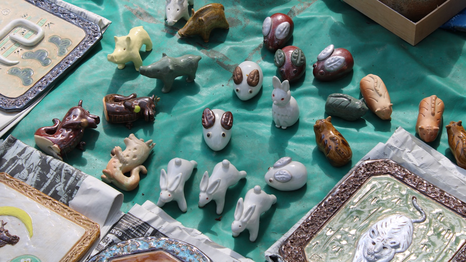 Small hand-painted ceramic zodiac animal figurines, including rabbits, mice, a dragon and a boar, arranged on a teal cloth at an outdoor stall.
