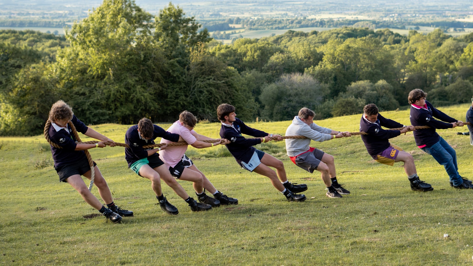 A team pulls hard on a rope during a tug-of-war contest on a grassy hillside, straining in the opposite direction from an unseen opposing team