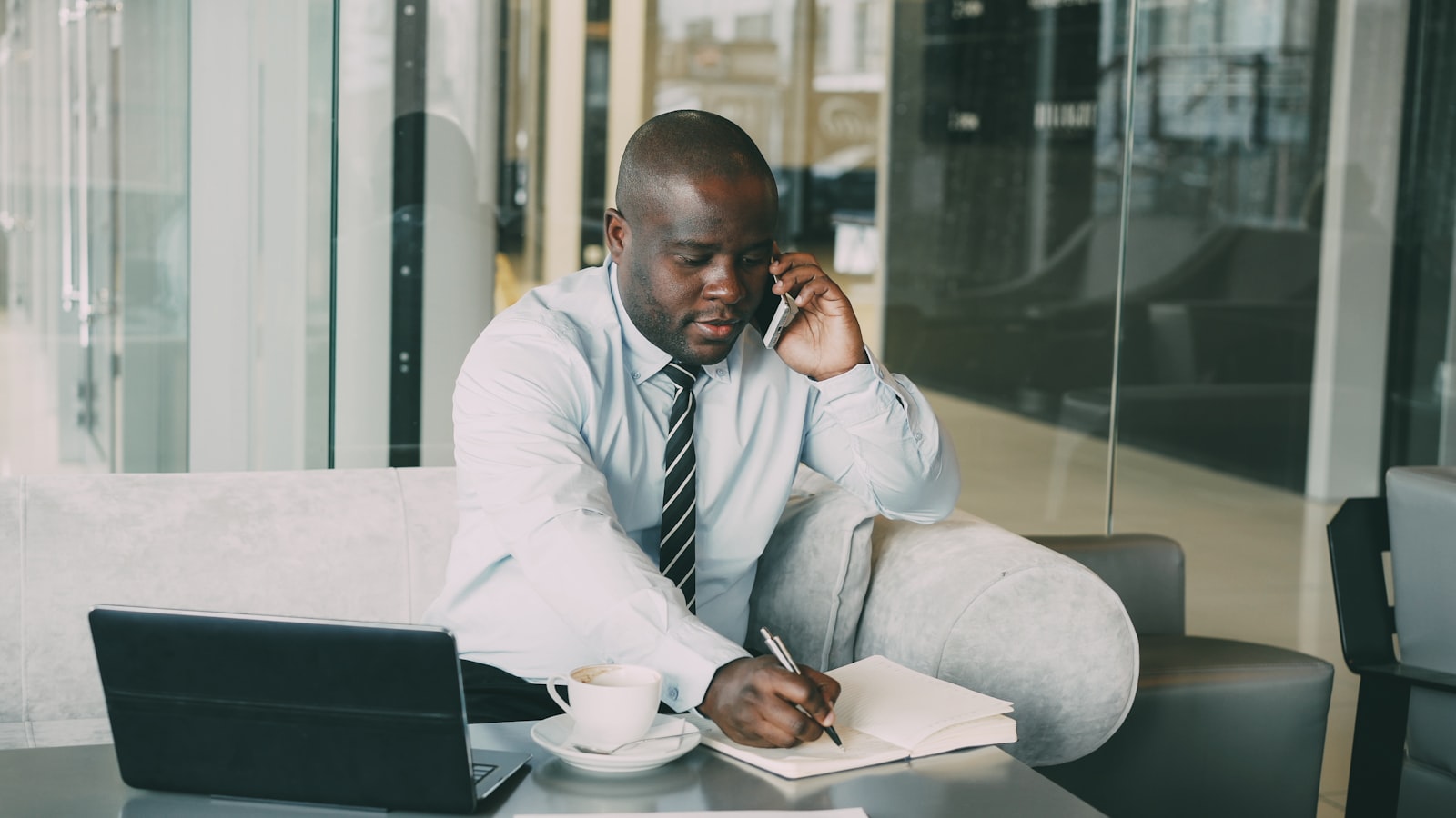 A man in a shirt and tie sits on a sofa on a phone call, writing in an open notebook with a laptop and coffee cup on the table in front of him.