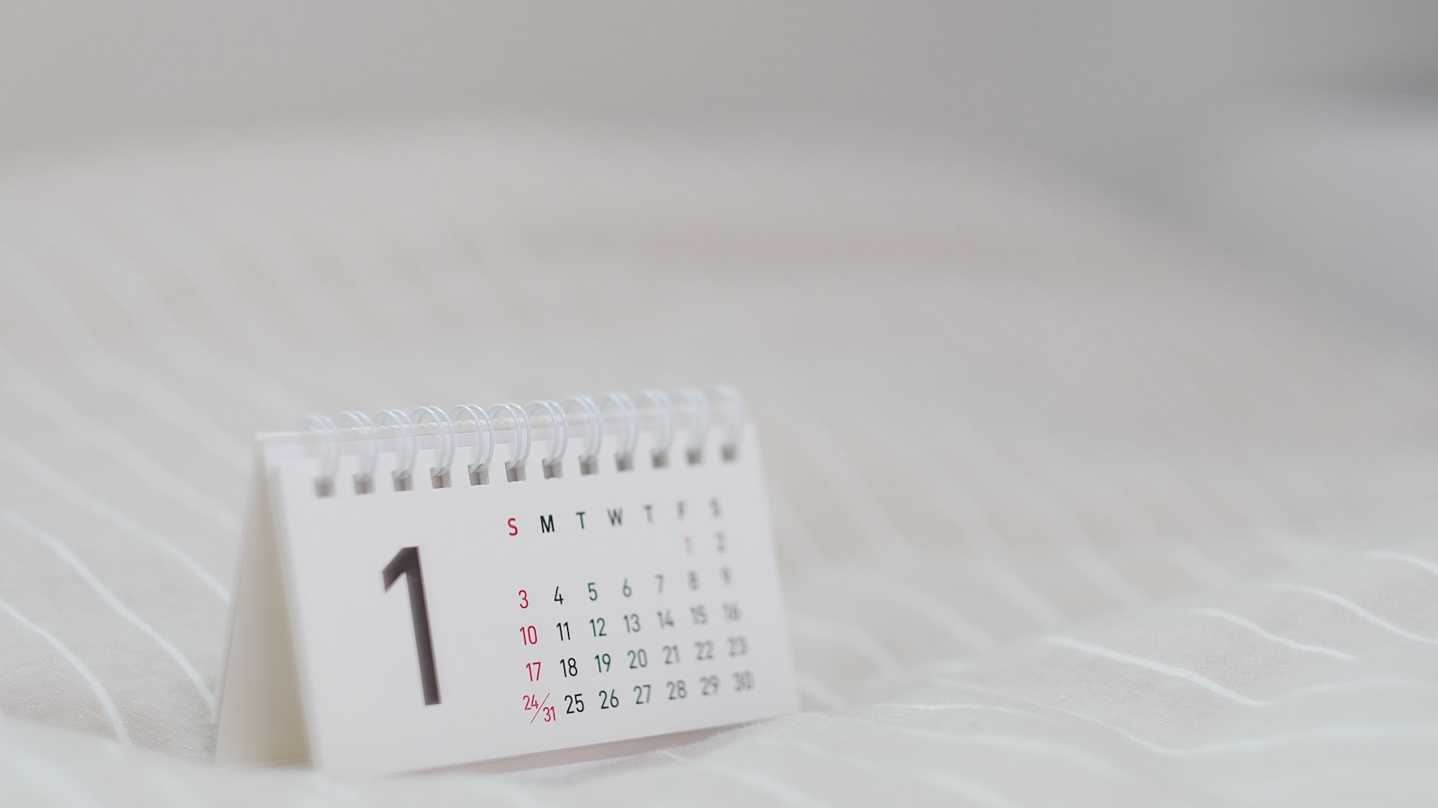 A small white spiral desk calendar showing the number 1, resting on soft white fabric
