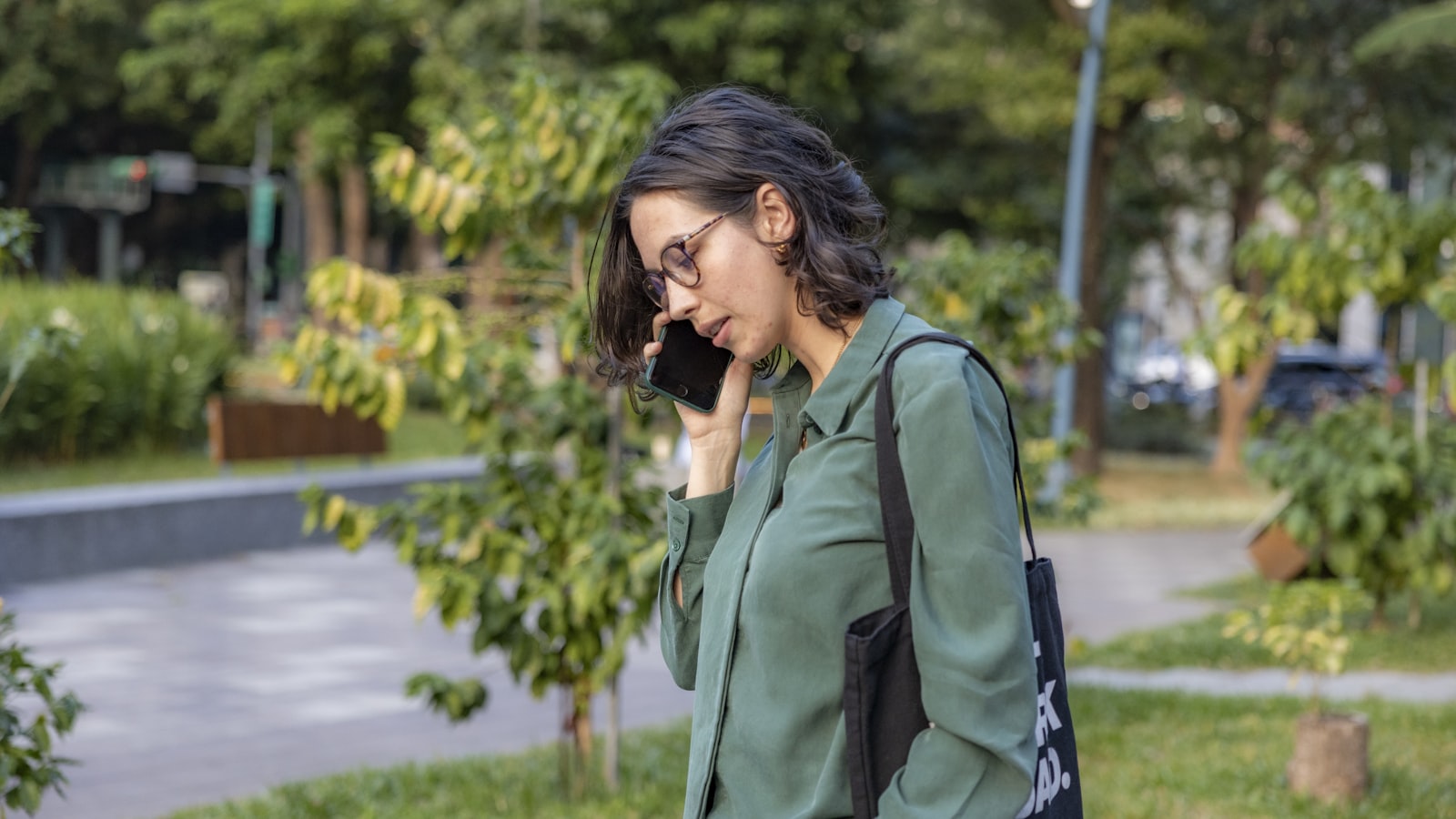 A woman standing outdoors in a park, smiling and gesturing while talking on her phone.